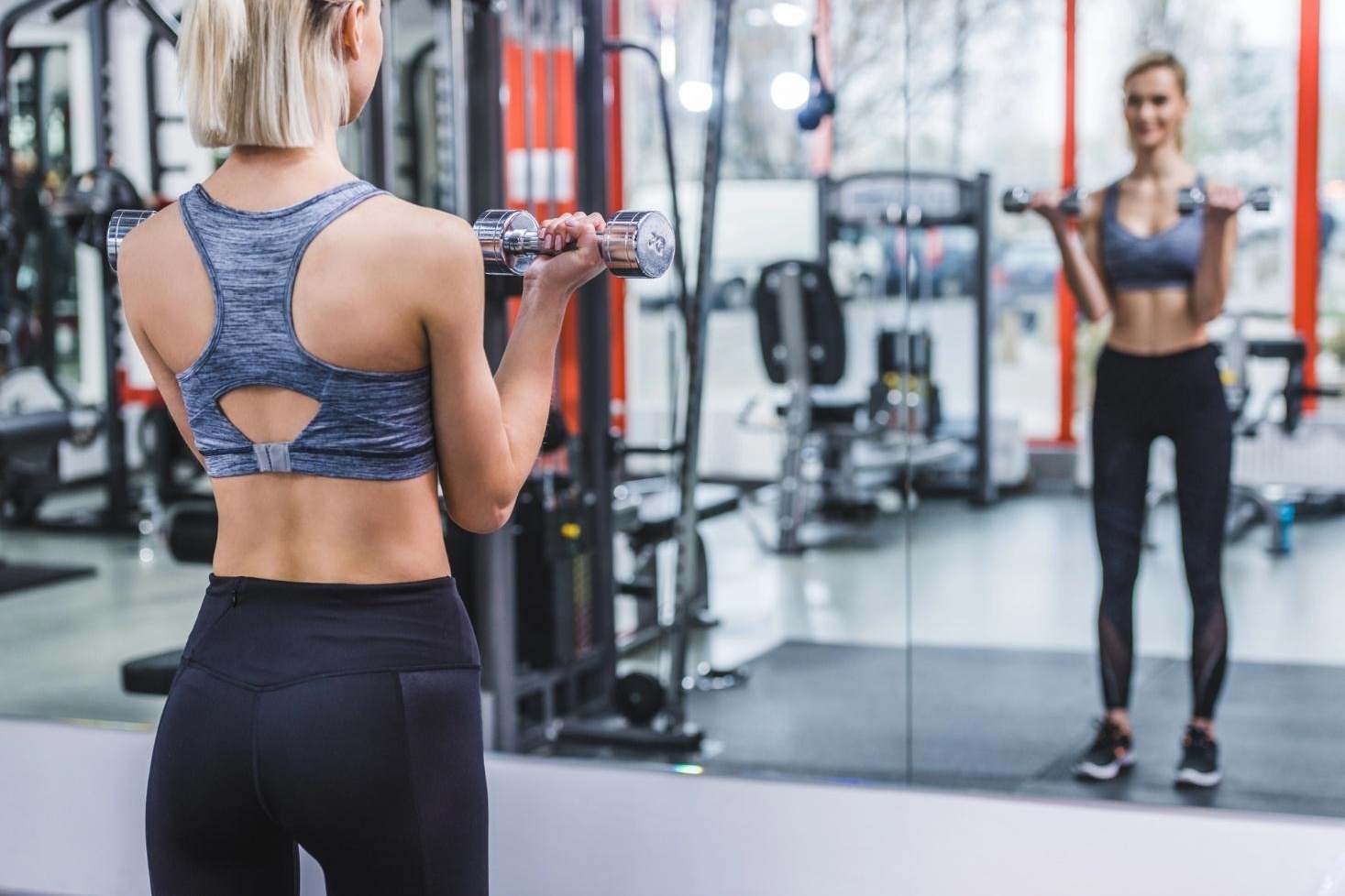 Athletic young woman working out with dumbbells in front of a mirror at the gym.