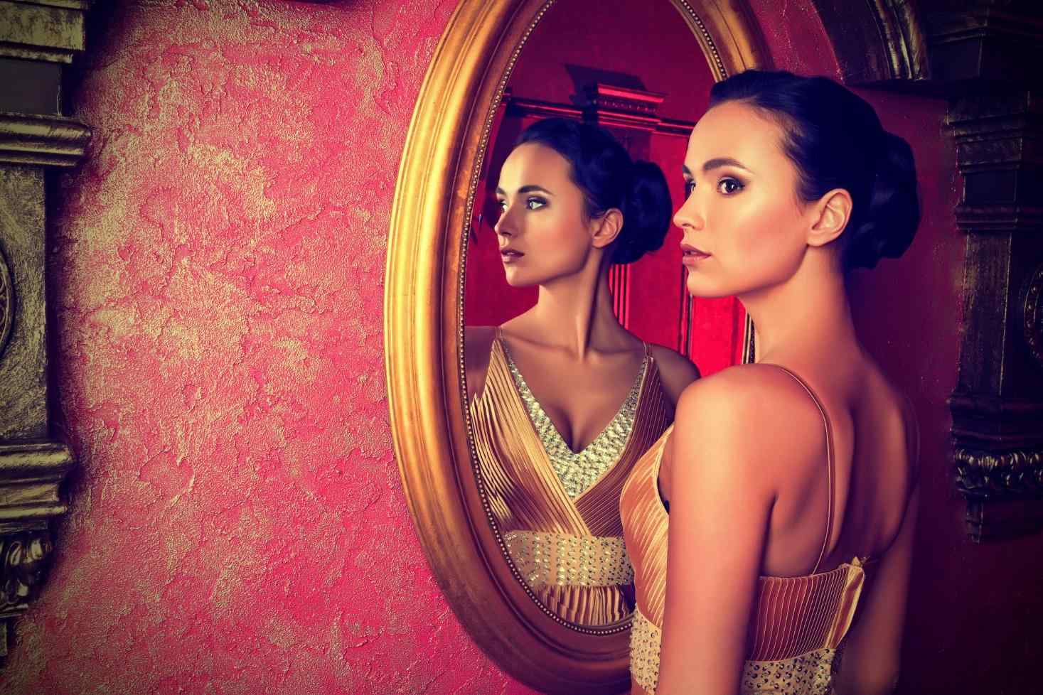 A beautiful woman stands before an antique mirror.